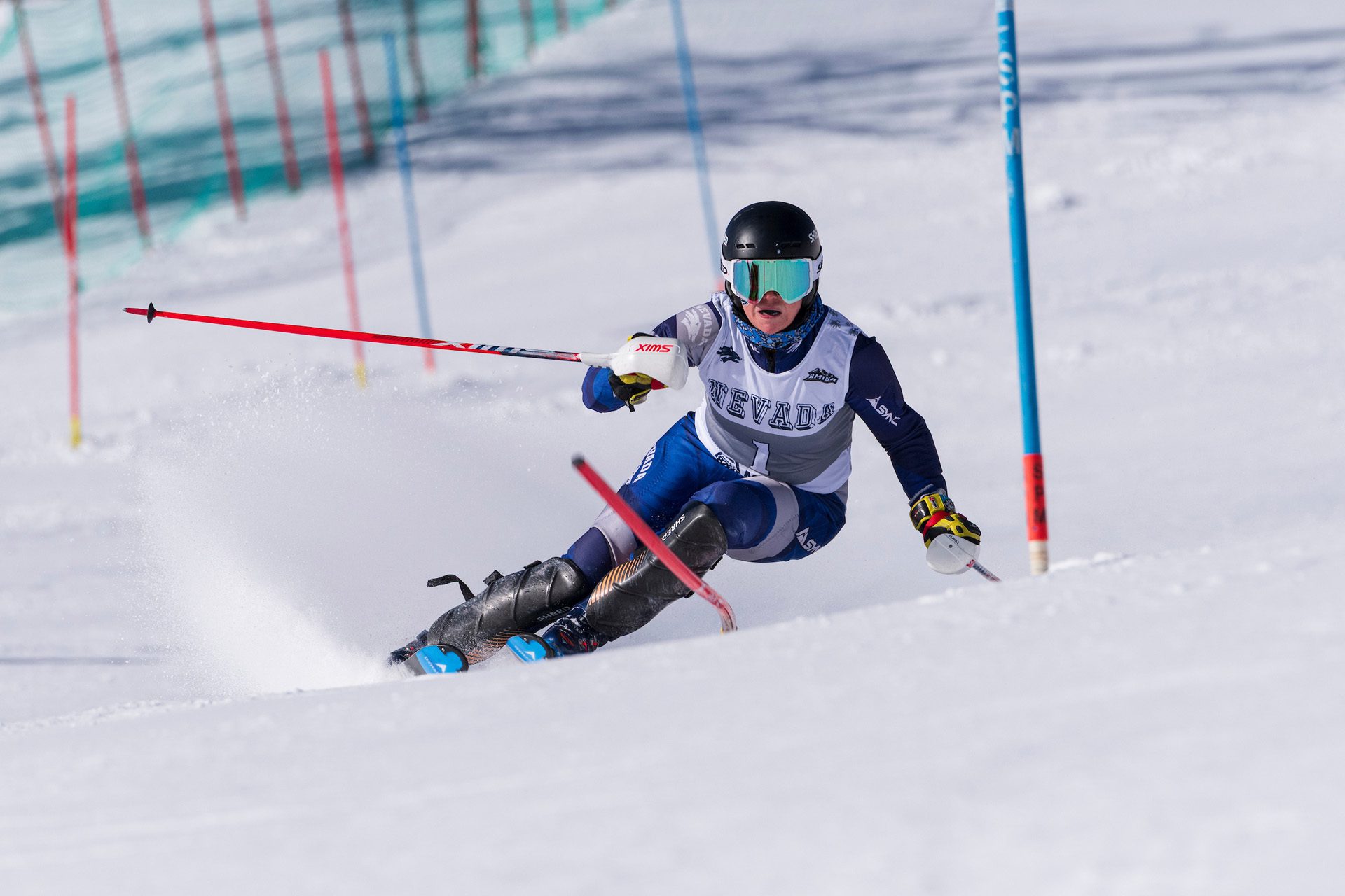 A skier in a racing suit and helmet competes in a slalom event, leaning into a turn on a snowy course marked by blue and red poles. Snow sprays up from their skis as they maneuver around a gate. Mt. Rose Ski Tahoe