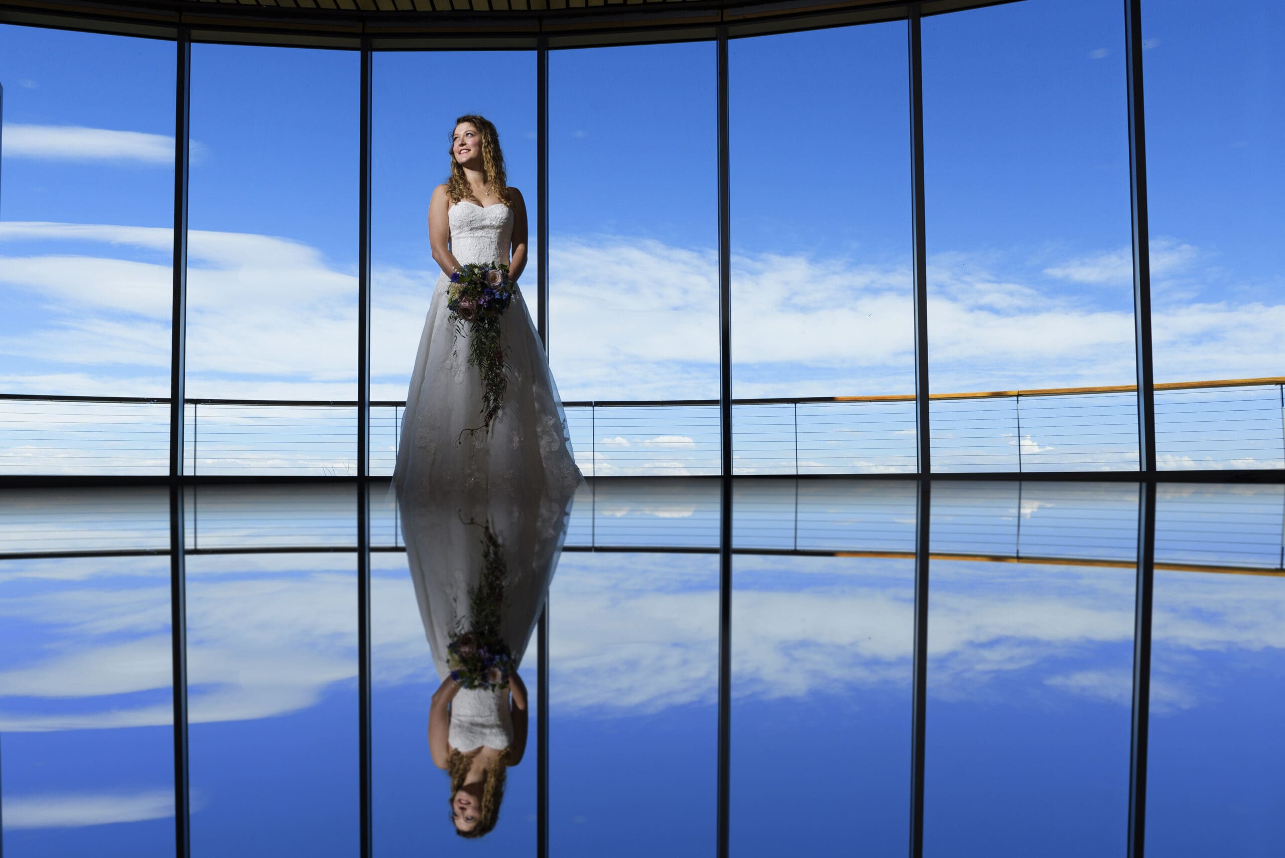 A bride in a white dress stands indoors before large windows, holding a bouquet. Blue sky and clouds are visible outside, and her reflection appears on the shiny floor. Mt. Rose Ski Tahoe
