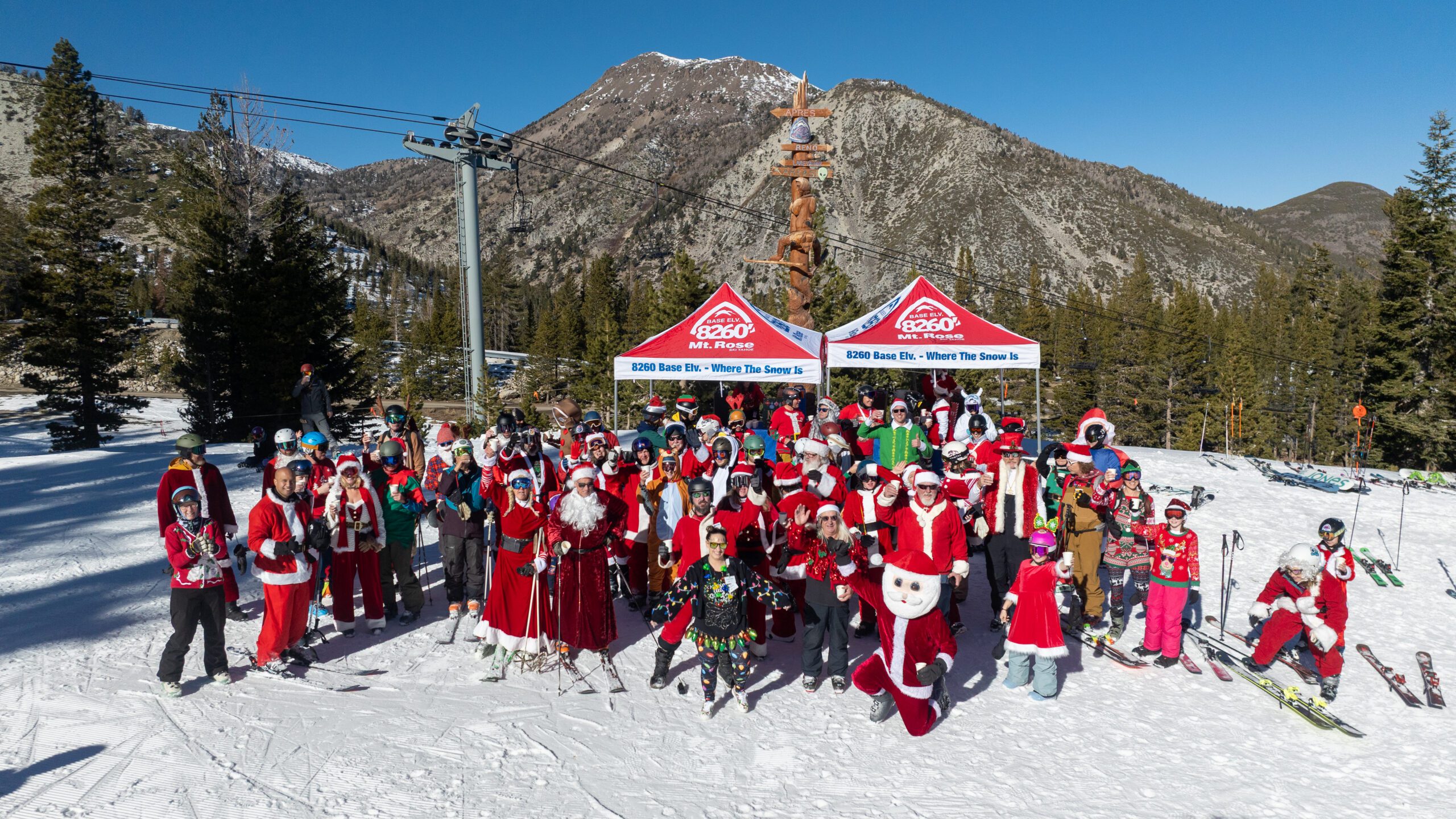 A large group of people dressed as Santa Claus and festive characters pose on a snowy mountain slope with ski gear, flanked by trees and mountains under a clear blue sky. Two red event tents are in the background. Mt. Rose Ski Tahoe