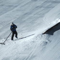 A person wearing winter gear uses a shovel to move snow near a ramp on a snow-covered surface, possibly preparing a ski or snowboard terrain park as part of their employment. Mt. Rose Ski Tahoe