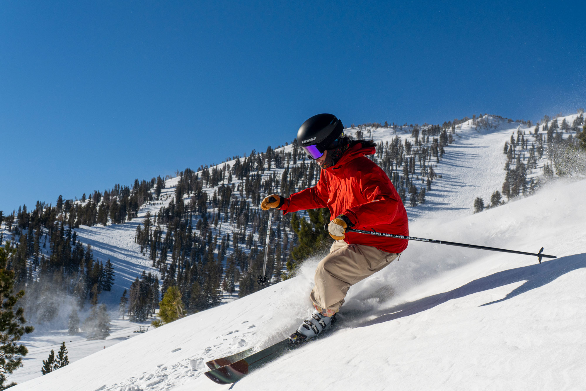 A skier in a red jacket, tan pants, and a black helmet skis downhill on a snow-covered slope with pine trees and clear blue sky in the background. Mt. Rose Ski Tahoe