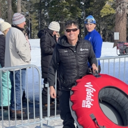 A man in sunglasses smiles while holding a red Wildslide snow tube. Other people in winter clothing stand nearby on the snowy ground with trees in the background. Mt. Rose Ski Tahoe