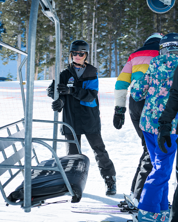 A Ski Tahoe lift operator in black winter gear assists two skiers, one in a colorful jacket and another in a floral jacket, as they prepare to board a ski lift on a snowy mountain. Mt. Rose Ski Tahoe