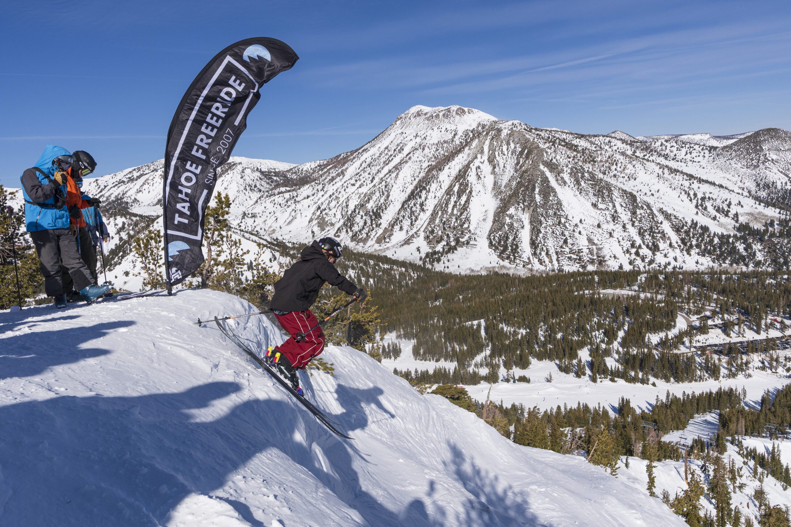 A skier in red pants launches off a snowy slope at a competition, with a “Tahoe Freeride” banner and mountains in the background under a clear blue sky. Mt. Rose Ski Tahoe