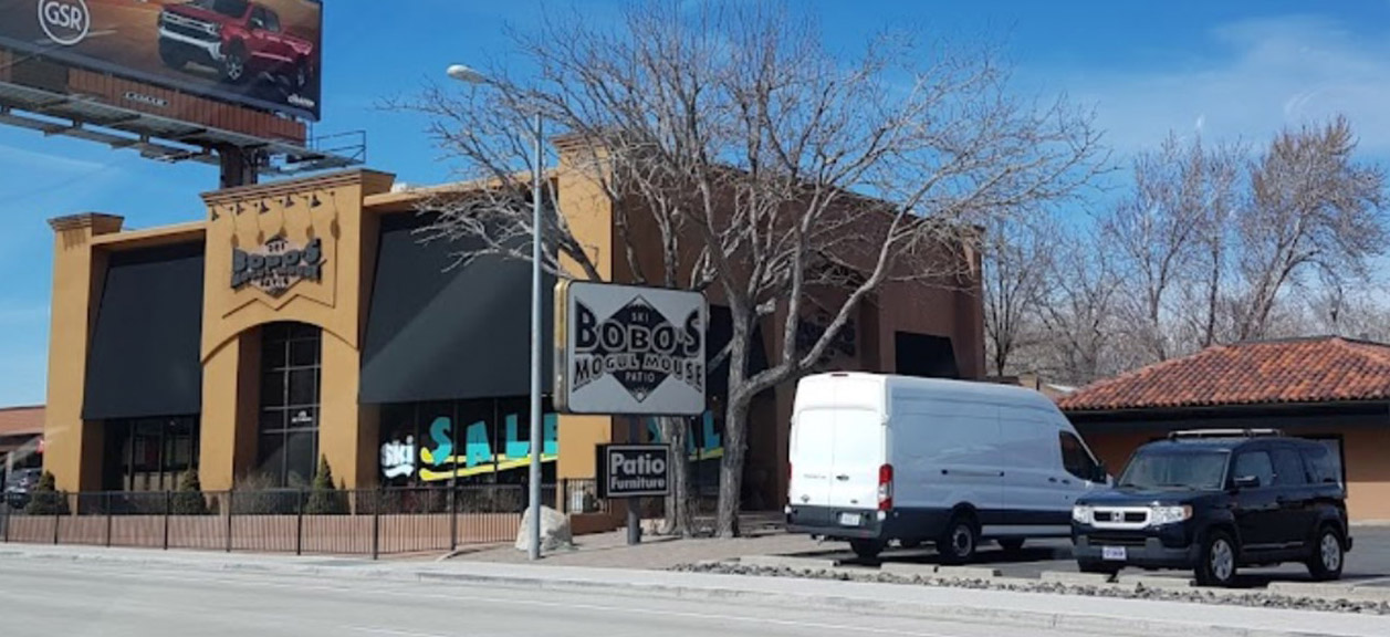 A restaurant named Bobos Mogollon House with a large sign, a white van, and a black SUV parked in front. Leafless trees and a billboard are visible in the background under a clear blue sky. Mt. Rose Ski Tahoe