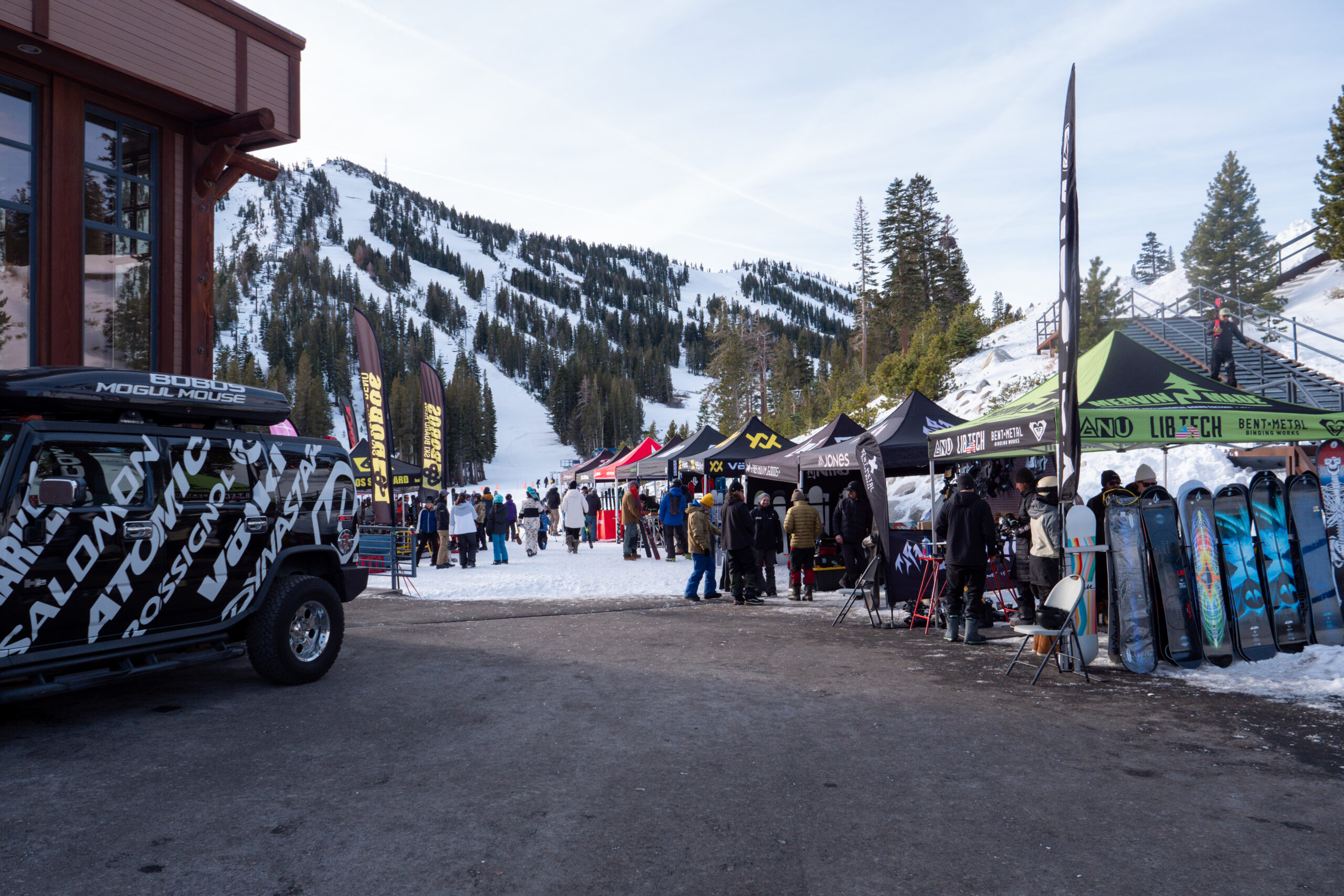 A snowy ski resort scene with people gathered near colorful tents, ski gear displays, and a branded vehicle. Snow-covered mountains and pine trees are visible in the background. Mt. Rose Ski Tahoe