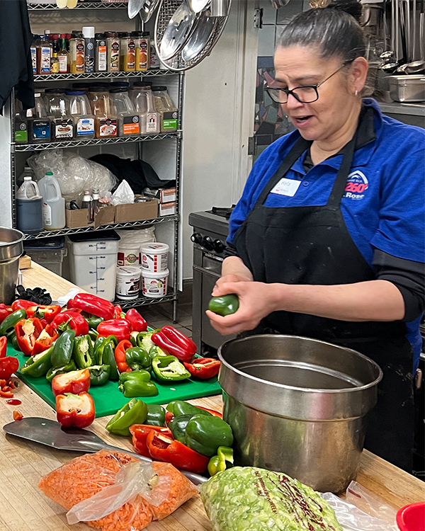 A kitchen worker wearing a blue uniform and black apron prepares green peppers on a cutting board in a Ski Tahoe commercial kitchen. Mt. Rose Ski Tahoe