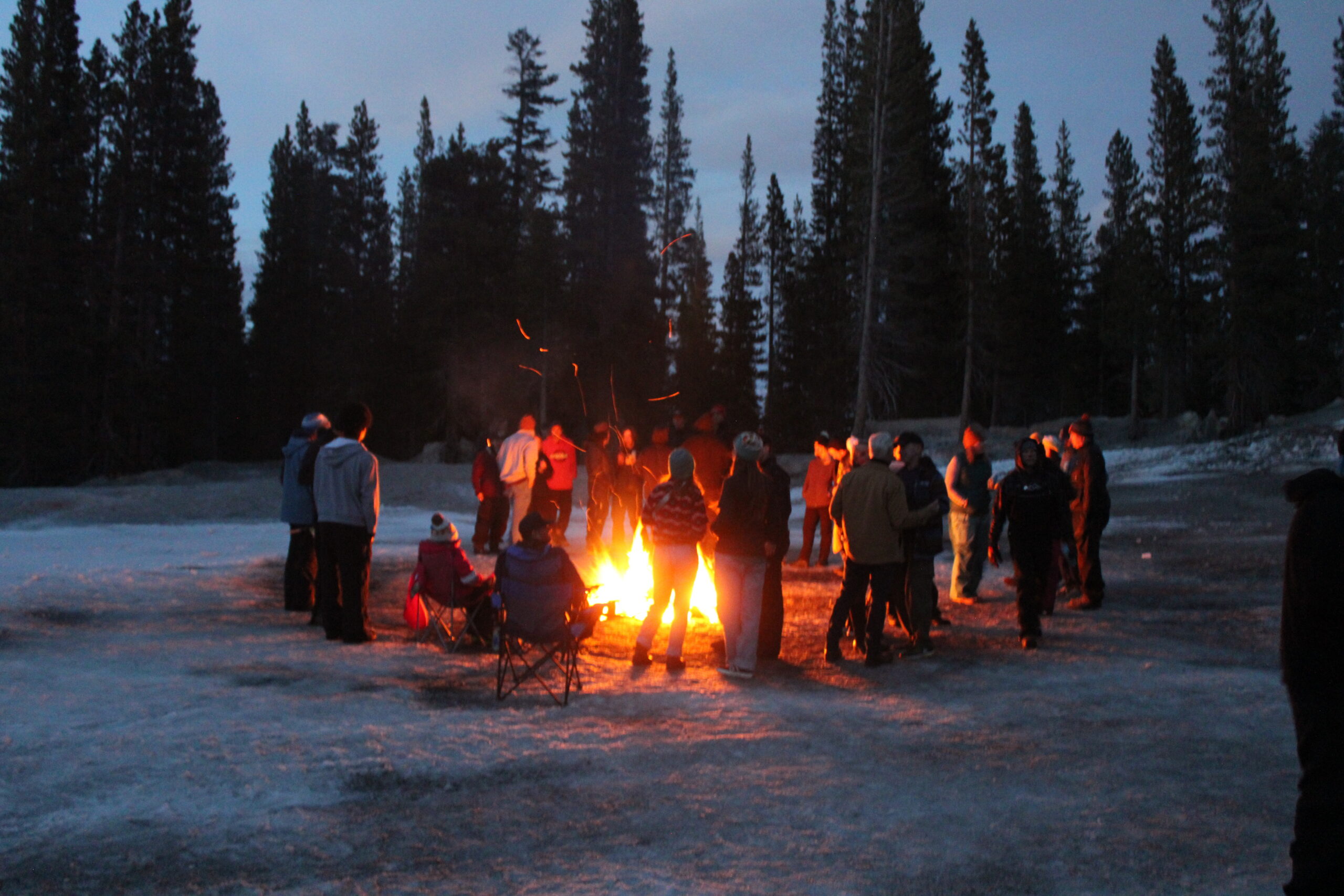 A group of people gathers around a campfire in a snowy, wooded area at dusk. Mt. Rose Ski Tahoe
