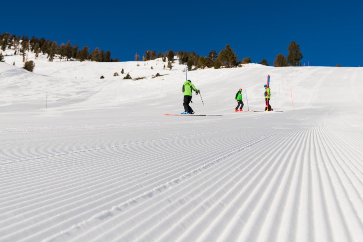 Three skiers stand on a groomed, corduroy-textured ski slope under a clear blue sky. Mt. Rose Ski Tahoe